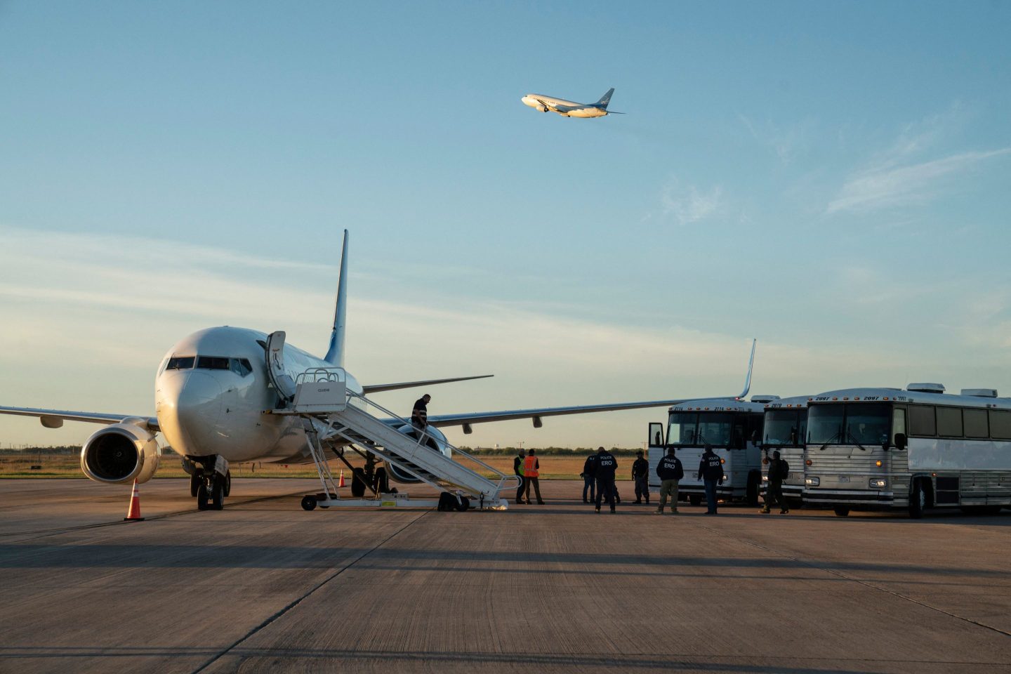 Buses transporting migrants to board the the first deportation flight of undocumented Venezuelans after a US-Venezuelan agreement are seen as another deportation plane headed to Central America is seen flying above them in Harlingen, Texas, on October 18, 2023.