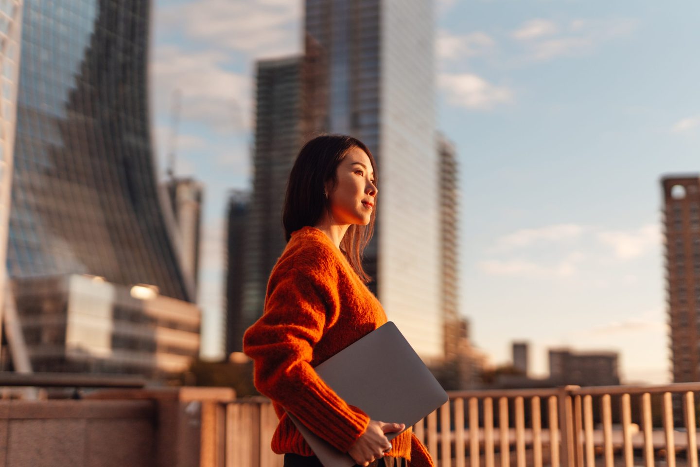 Confident young Asian businesswoman with laptop walking in the city against skyscraper.
