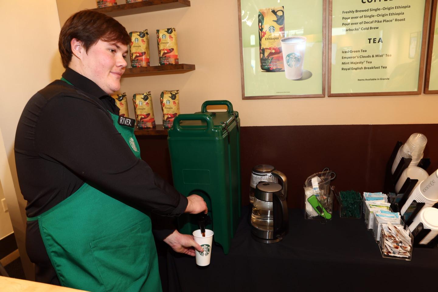 A barista with a black shirt and green apron fills up a cup with coffee.