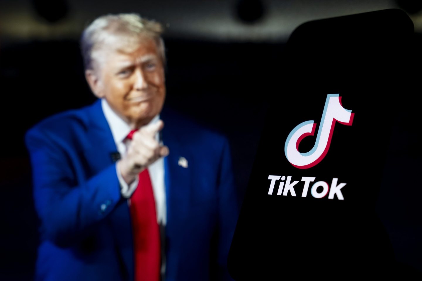 The logo of TikTok is displayed on mobile phone screen in front of a computer screen displaying United States President Donald Trump