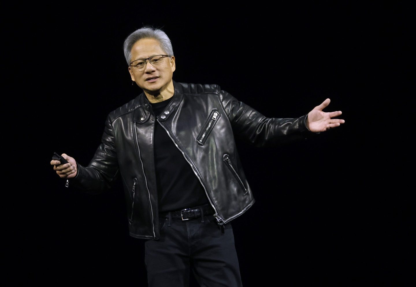 Nvidia CEO Jensen Huang in a black jacket on stage against a black background with his arms outstretched.