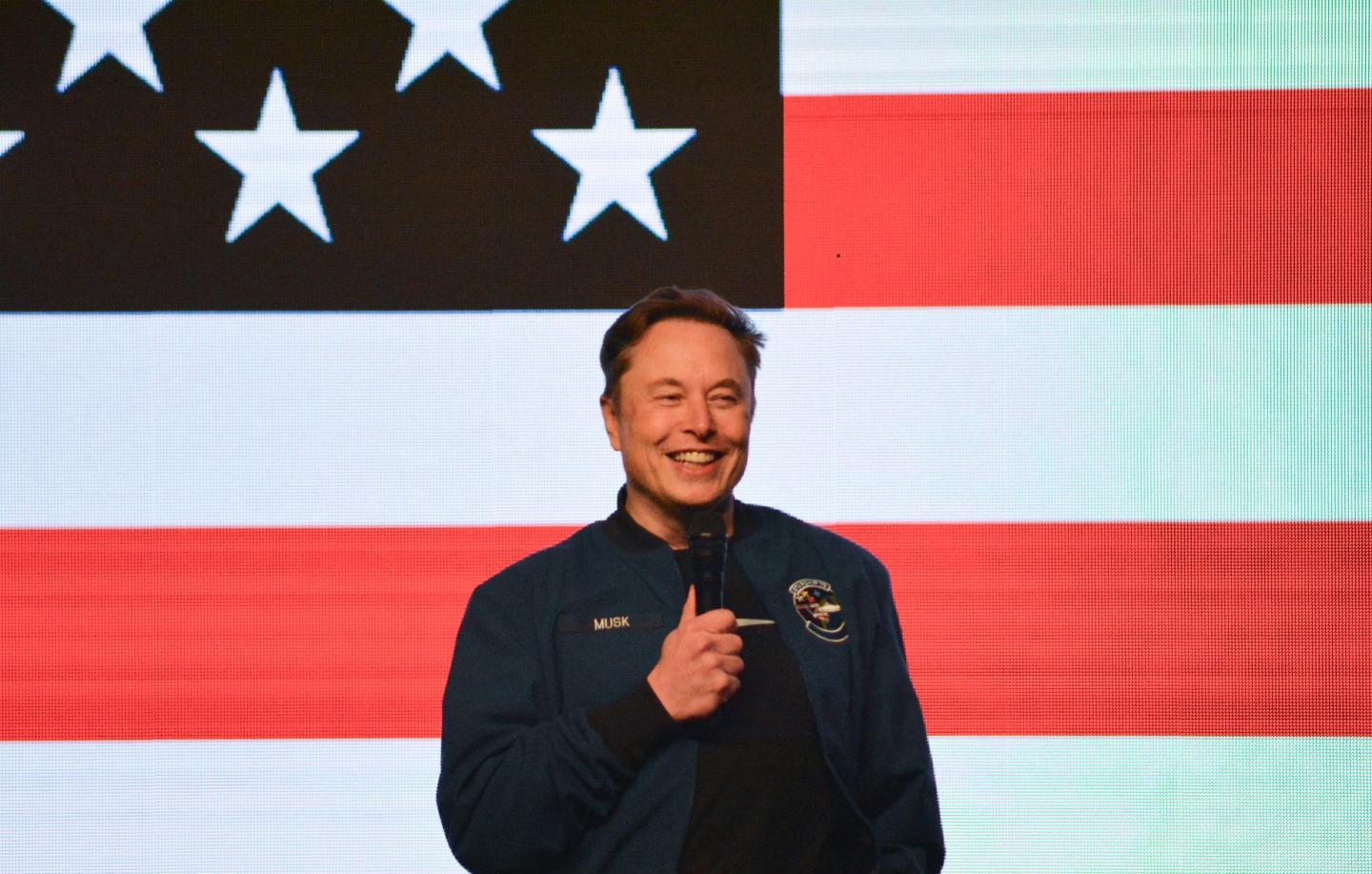 Elon Musk holds a microphone on stage in front of an American flag