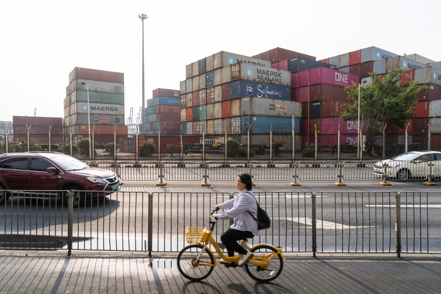 A woman rides a bicycle across the street from high stacks of large shipping containers.
