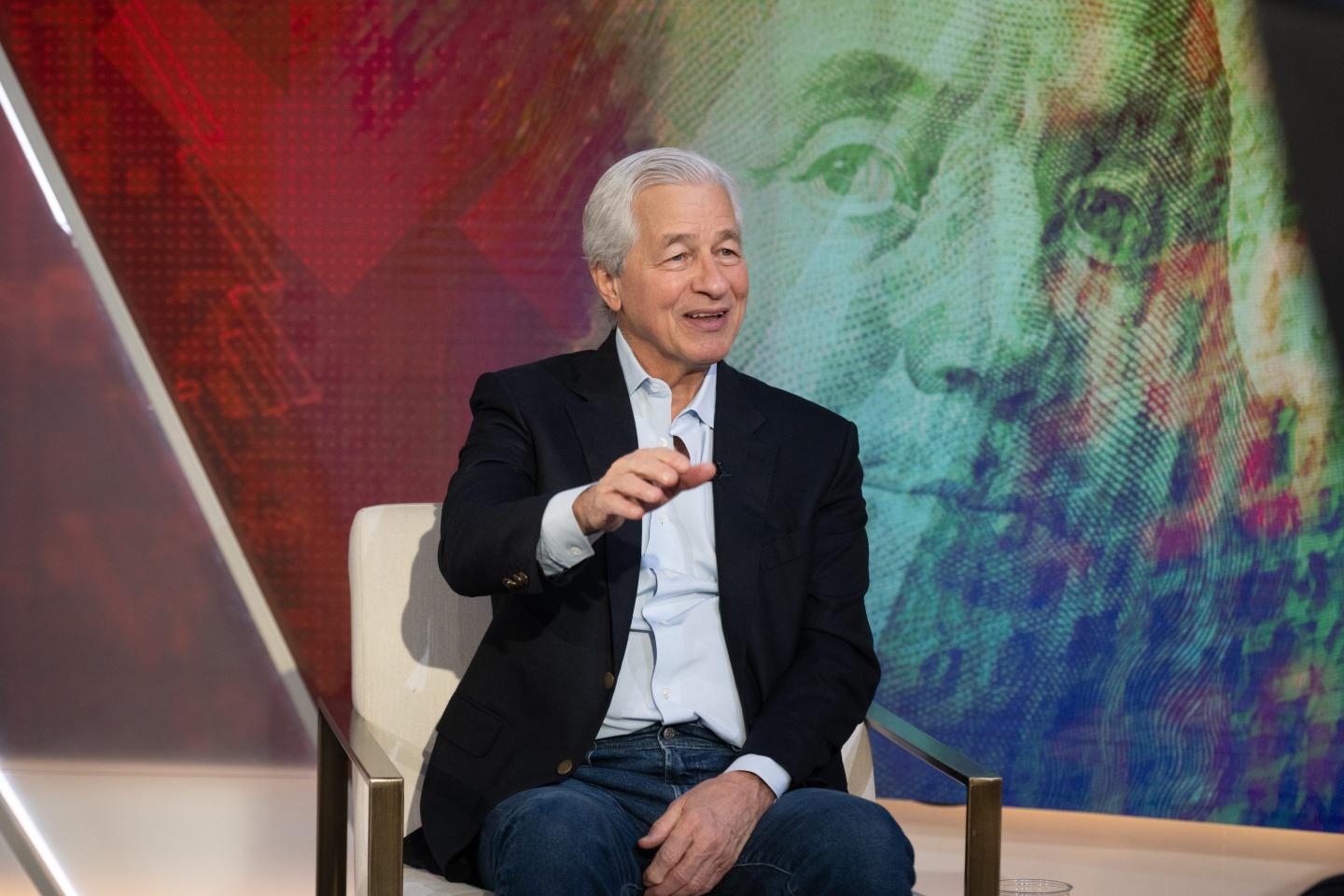Jamie Dimon smiles and lifts his right hand as he talks seated in front of a background with an image of Benjamin Franklin.