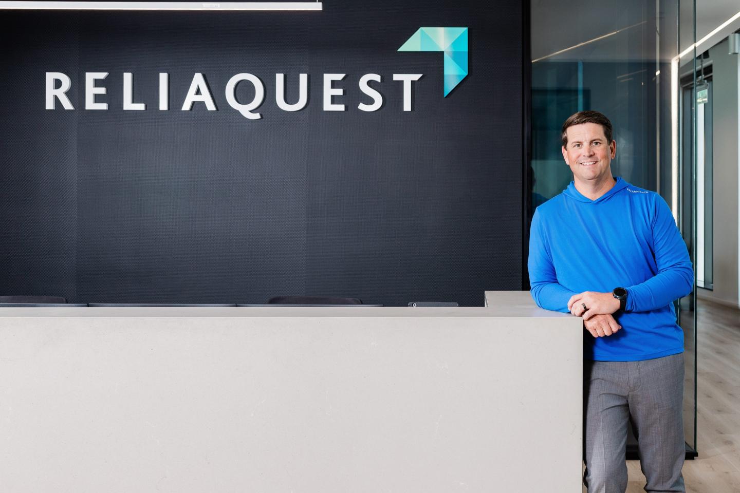 ReliaQuest CEO Brian Murphy leaning against a desk, with the company logo above it