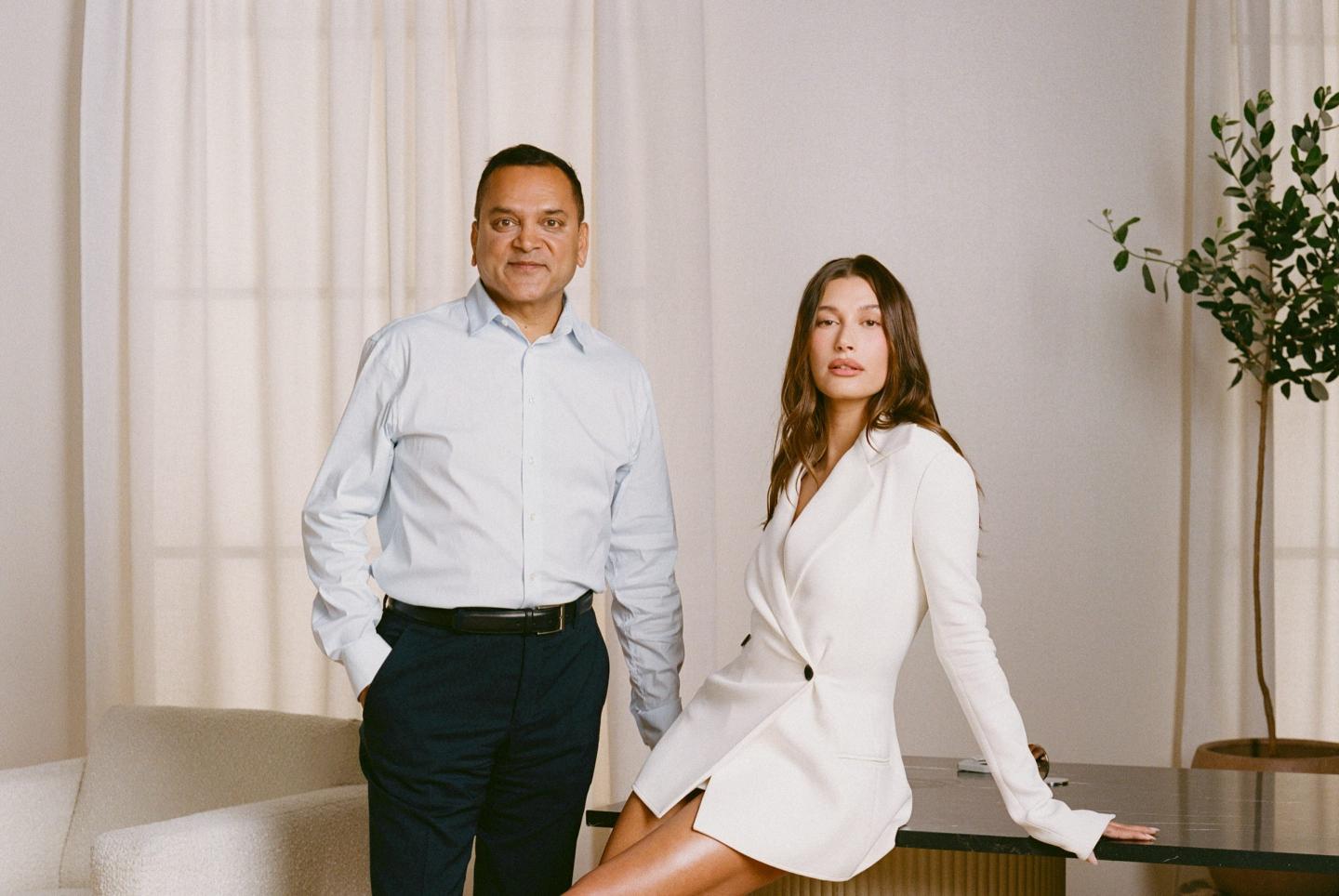 Tarang Amin and Hailey Bieber lean on a glass table and look forward.