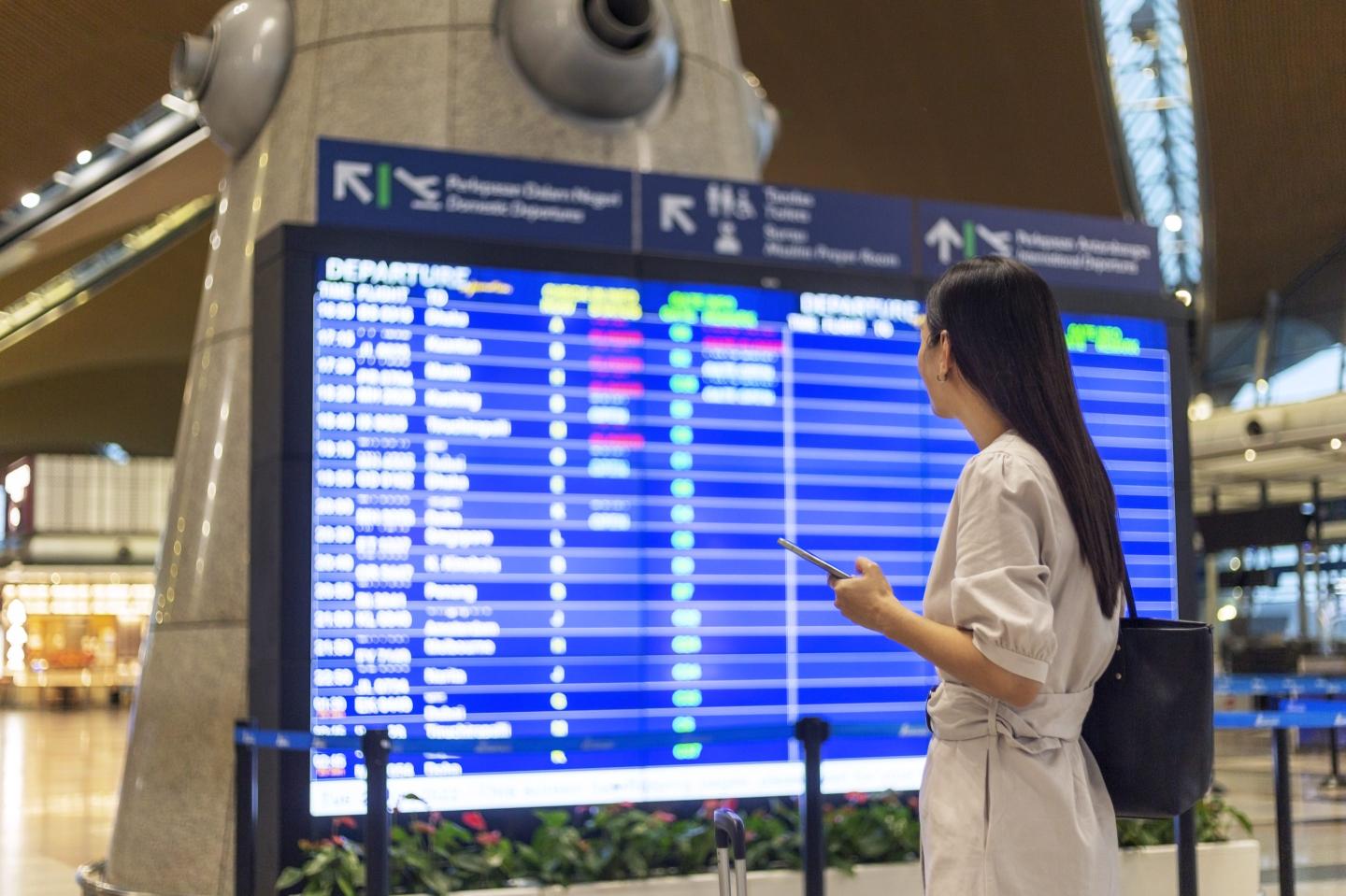 woman with luggage and smart phone checking flight schedule near departure board in airport