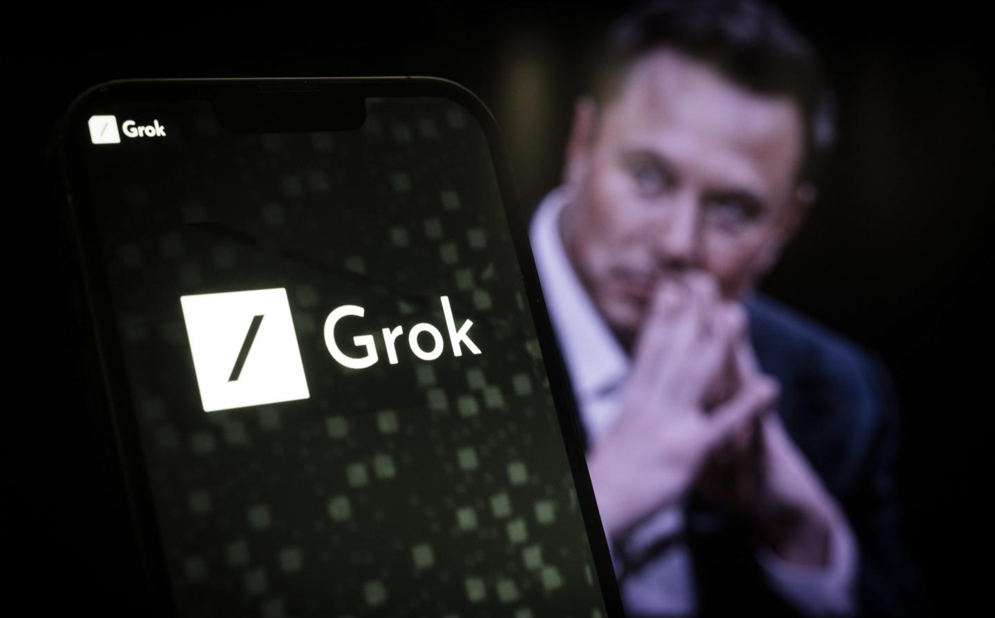 Elon Musk folds his hands behind a logo of xAI and Grok