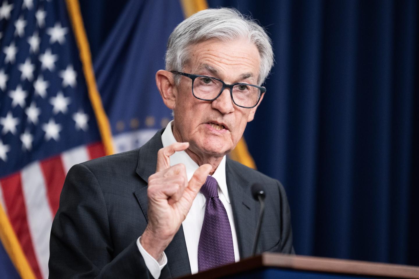 Fed Chair Jerome Powell creates a space between his thumb and the index finger on his right hand as he speaks at a podium with an American flag in the background.