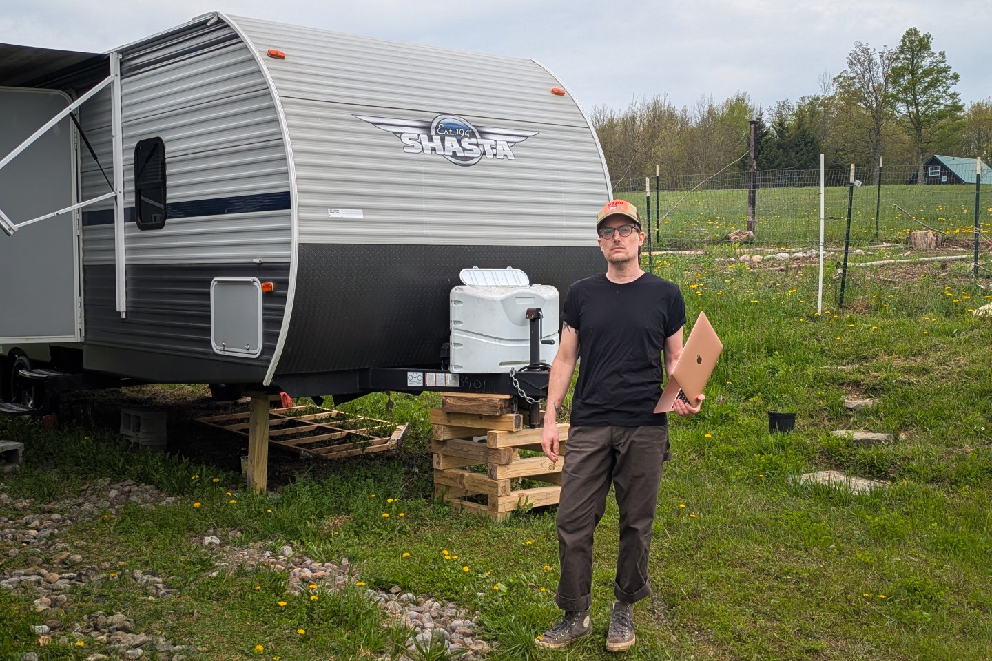 Shawn K next to his RV trailer, wearing a hat and holding a laptop.