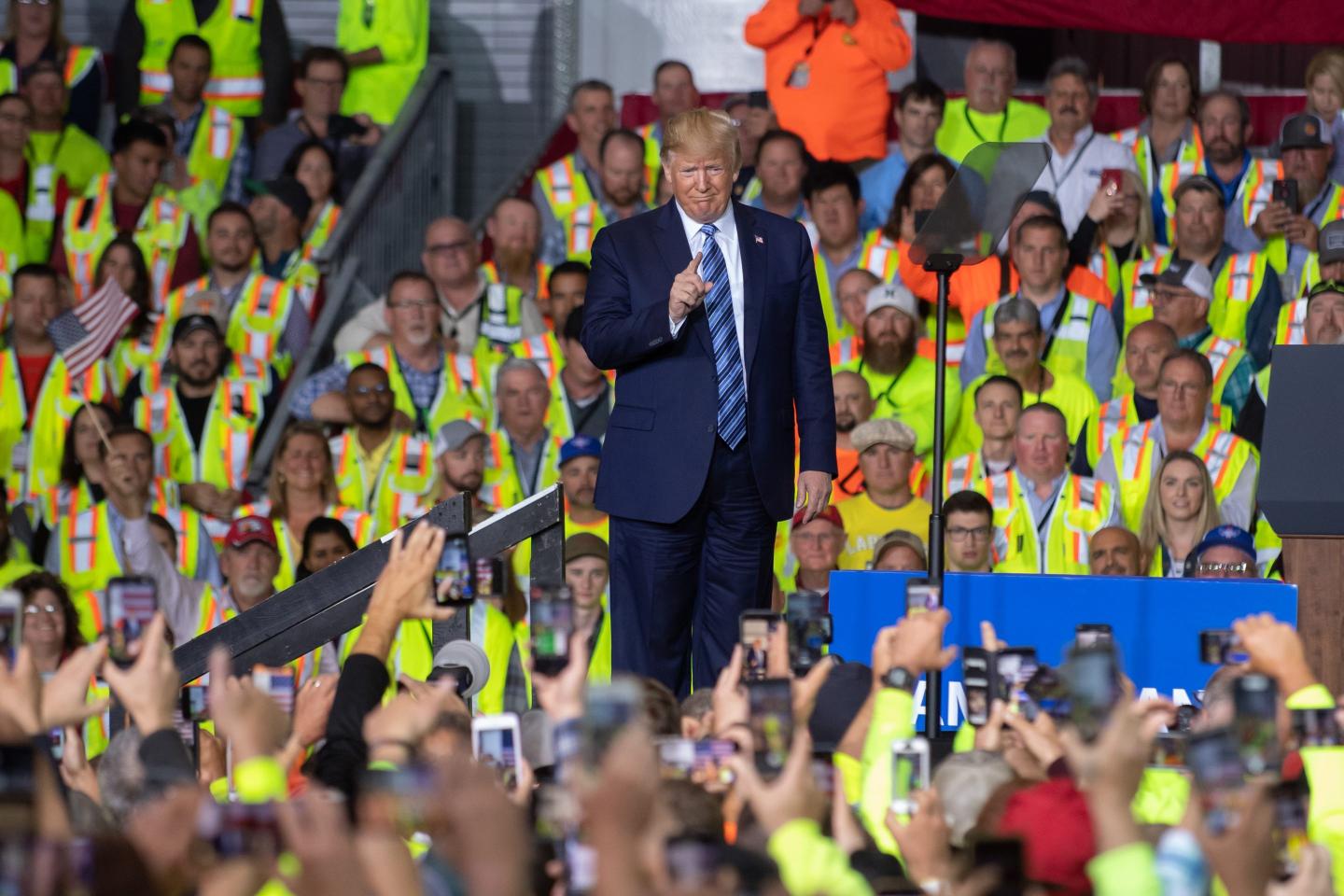 Donald Trump points his index finger at cheering workers dressed in bright green construction vests with orange and grey stripes.