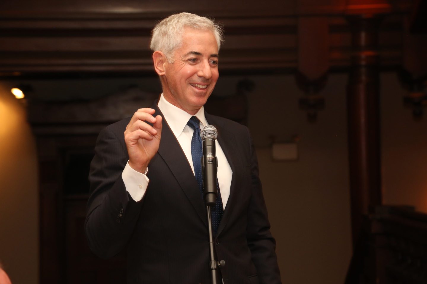 Bill Ackman smiles and raises his right hand as he speaks in front of a microphone.