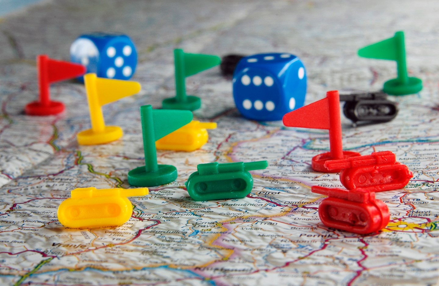 An image of small plastic colored flags and miniature tanks, as might be used in a war strategy board game, arrayed on top of a map with a pair of blue dice in the background.