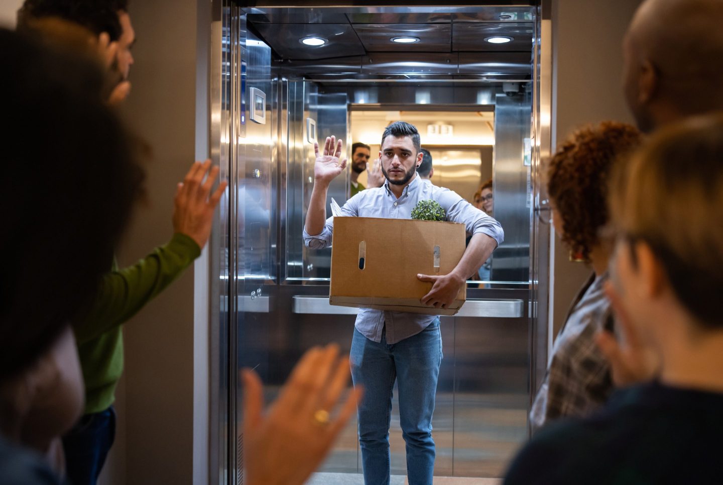 A man in an elevator holding a box of his office things waves goodbye to his now ex-coworkers