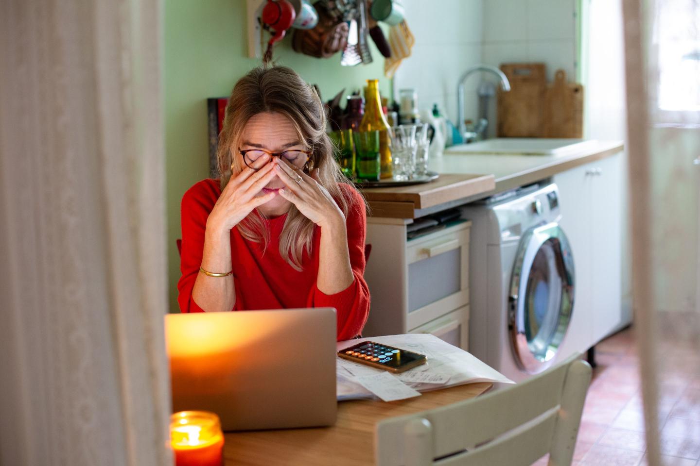 A woman, looking stressed and exhausted, rubs her eyes beneath her glasses as she sits at a kitchen table with her laptop open in front of her.