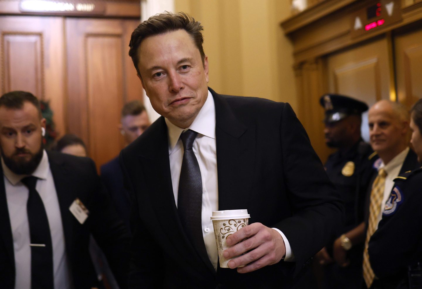 Elon Musk, wearing a suit, carries a paper cup in his hand.