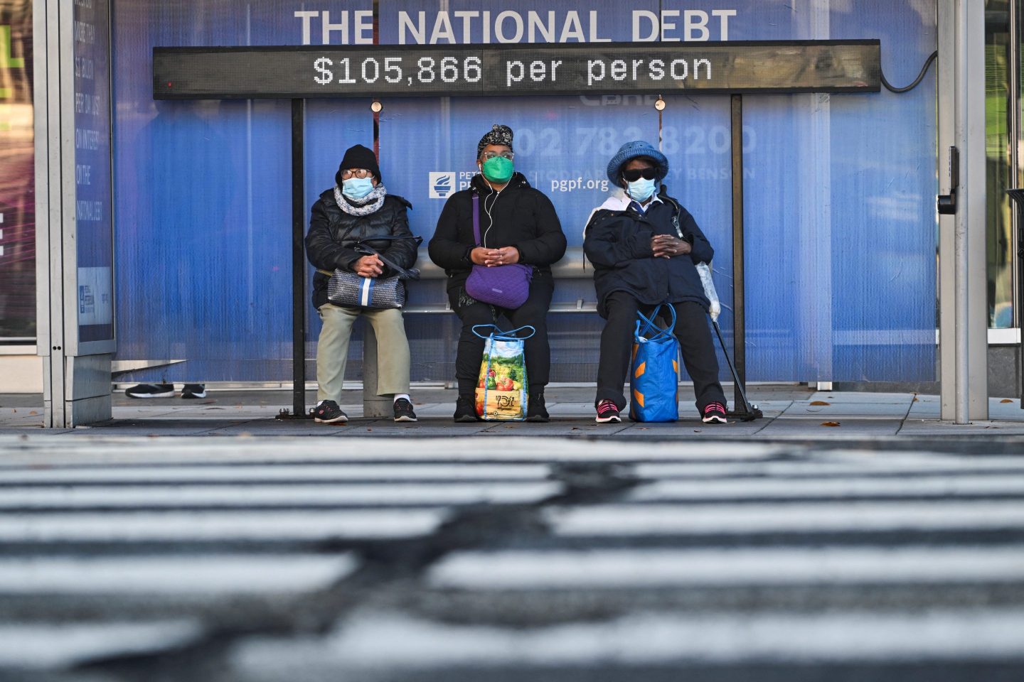 Commuters wait at a bus stop where there is a National Debt Clock billboard-sized running total display that shows the United States gross national debt as $105,866 per person in Washington, DC, on December 30, 2024.