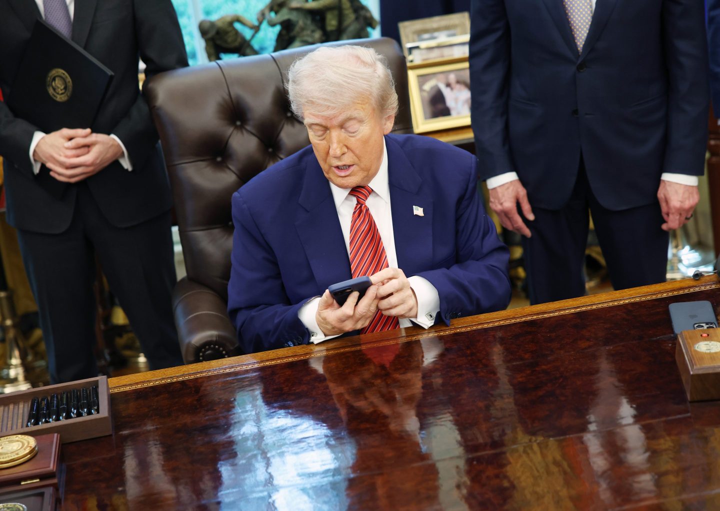 Donald Trump, sitting at his desk in the Oval Office, looks down at his phone.