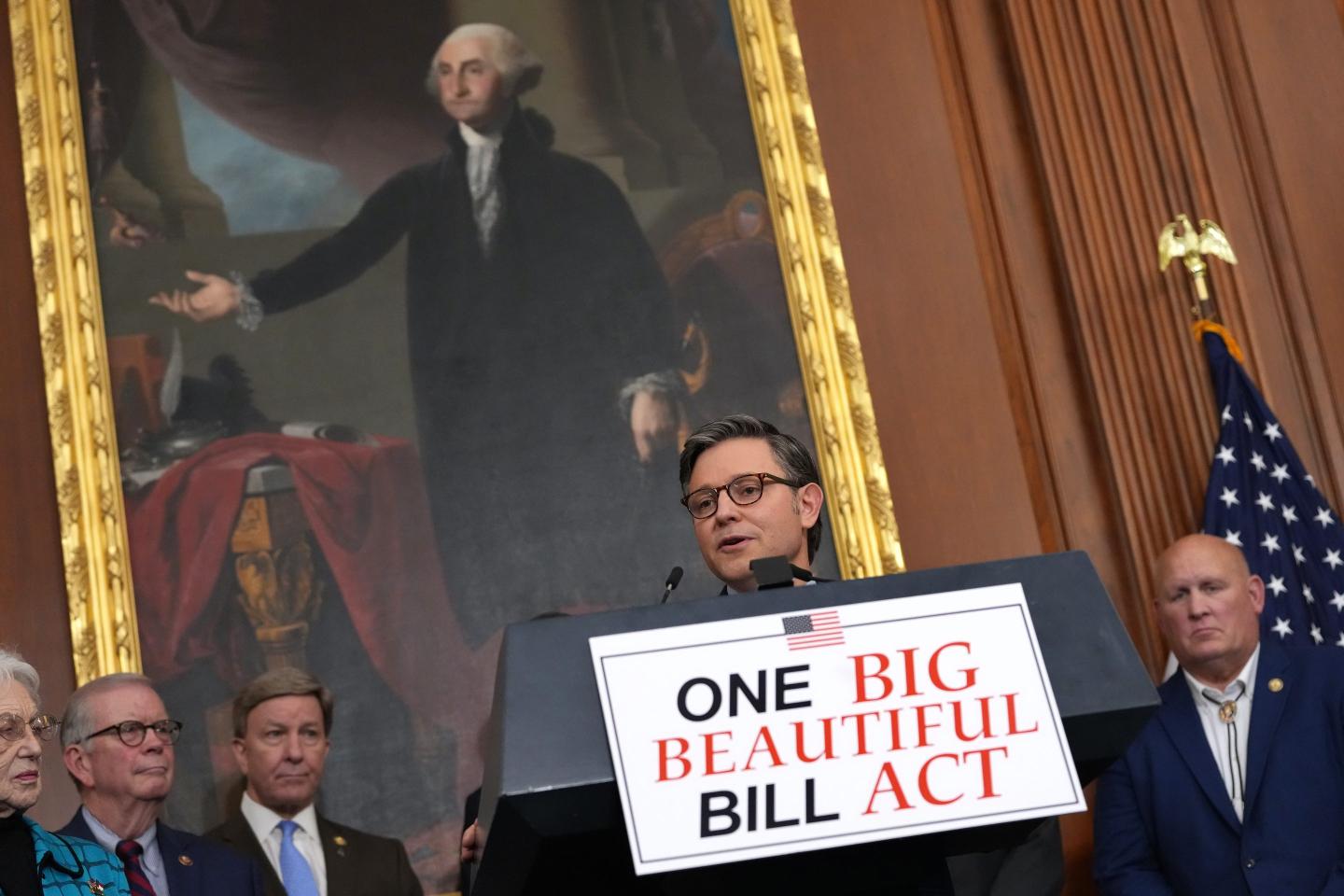 Speaker of the House Mike Johnson speaks at a podium with a placard reading "ONE BIG BEAUTIFUL BILL ACT." A portrait of George Washington hangs in the background.