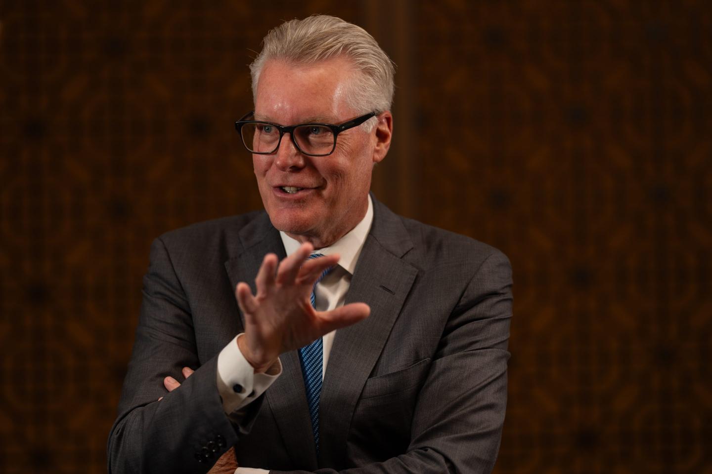 A man with graying hair and glasses speaks to someone off-camera.