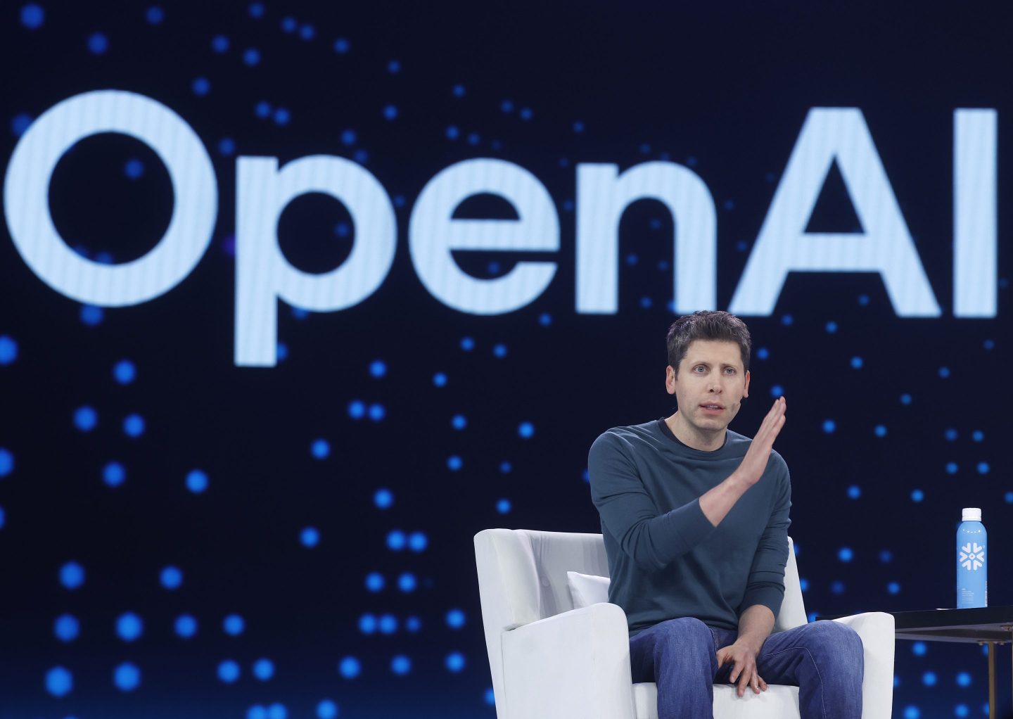 A photo of OpenAI CEO Sam Altman sitting on stage in front of the word OpenAI projected on a big screen behind him making a gesture with his right hand that looks like a line on a graph sloping steeply up to the right.