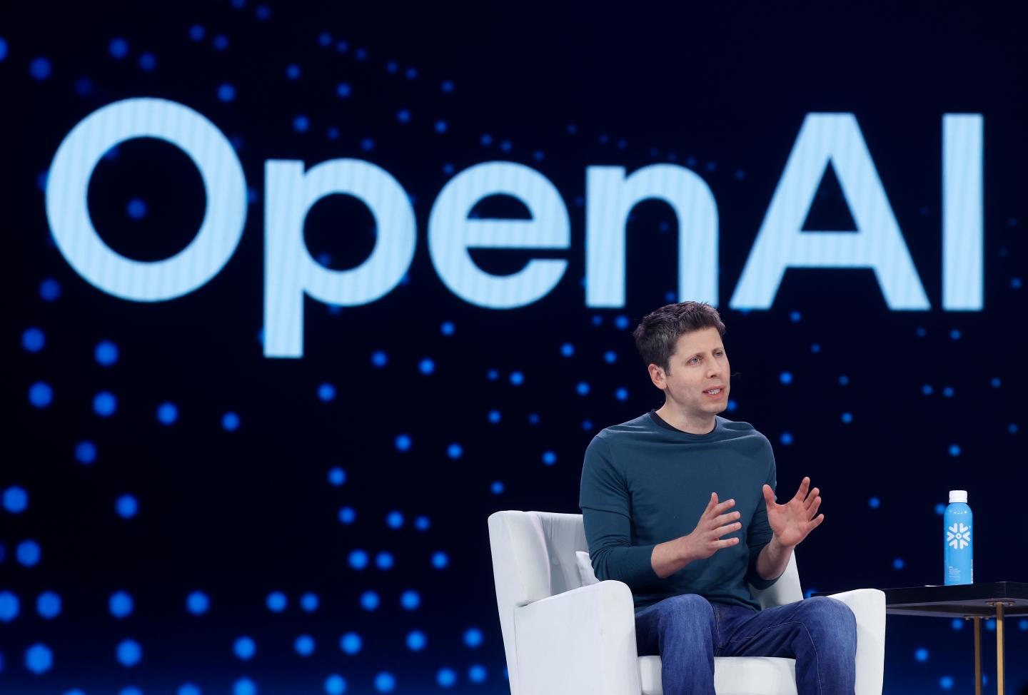 Sam Altman sitting on a chair in front of the OpenAI logo.