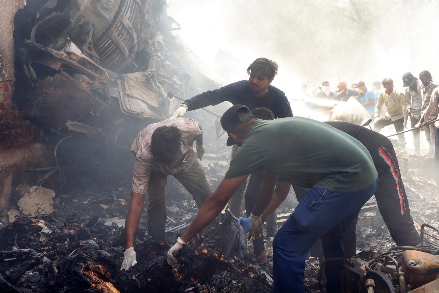 Three people pick up wreckage next to a crashed jet, surrounded by smoke.