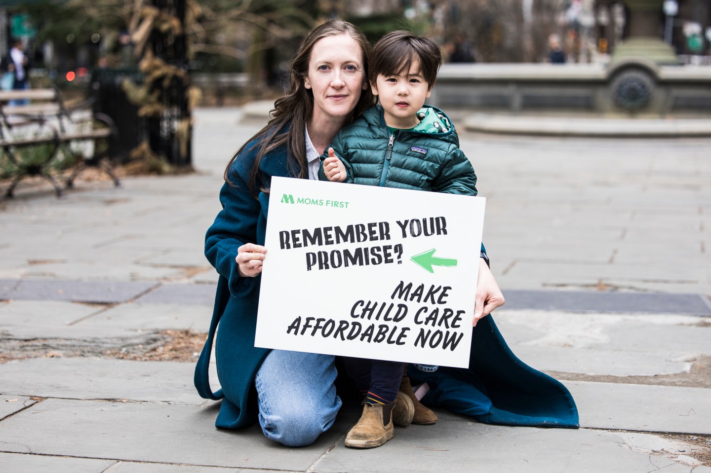 Mother and child holding a Moms First sign, crouching on a city sidewalk