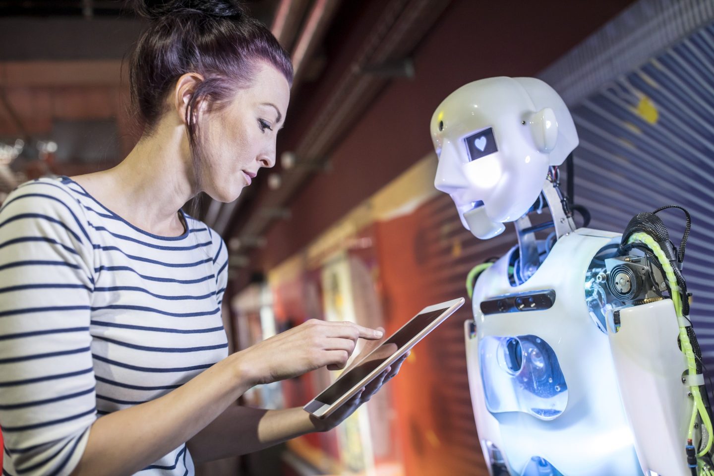 A woman wearing a striped top consults at a digital tablet as she stands in front of a humanoid robot.