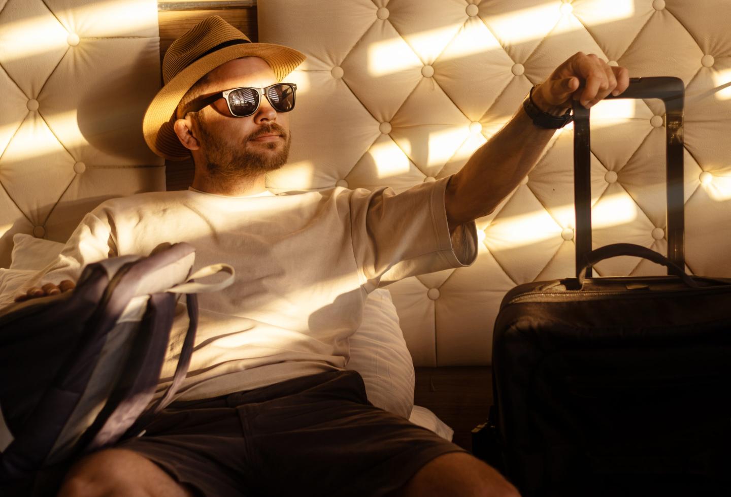 man wearing a hat and sunglasses lying on the bed in the hotel and holding a bag