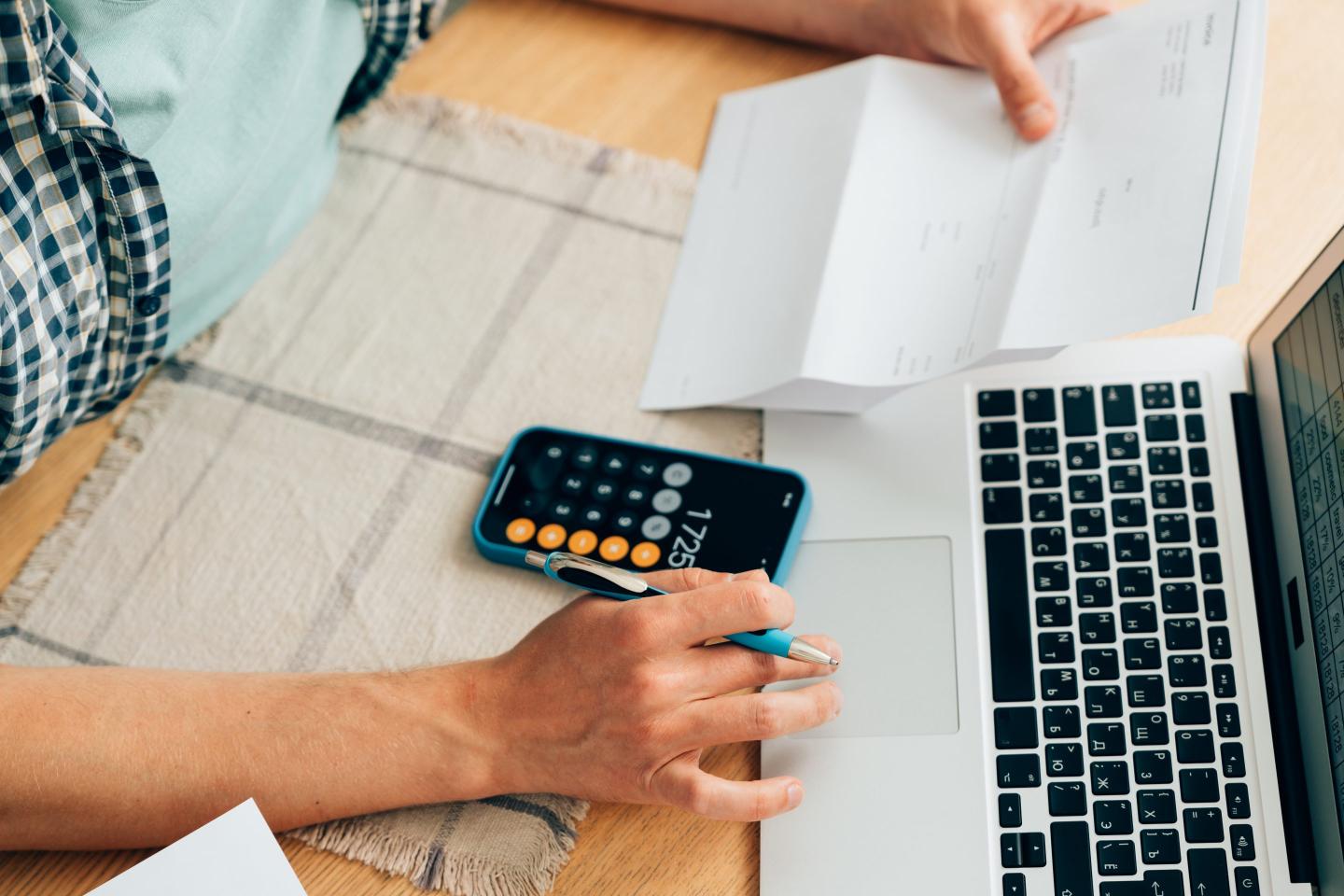 Unrecognizable person counting expenses on calculator in mobile phone app, checking her energy bills at home. Budget management, price increases, inflation, rise in price of products and utilities. View from above, managing home finances