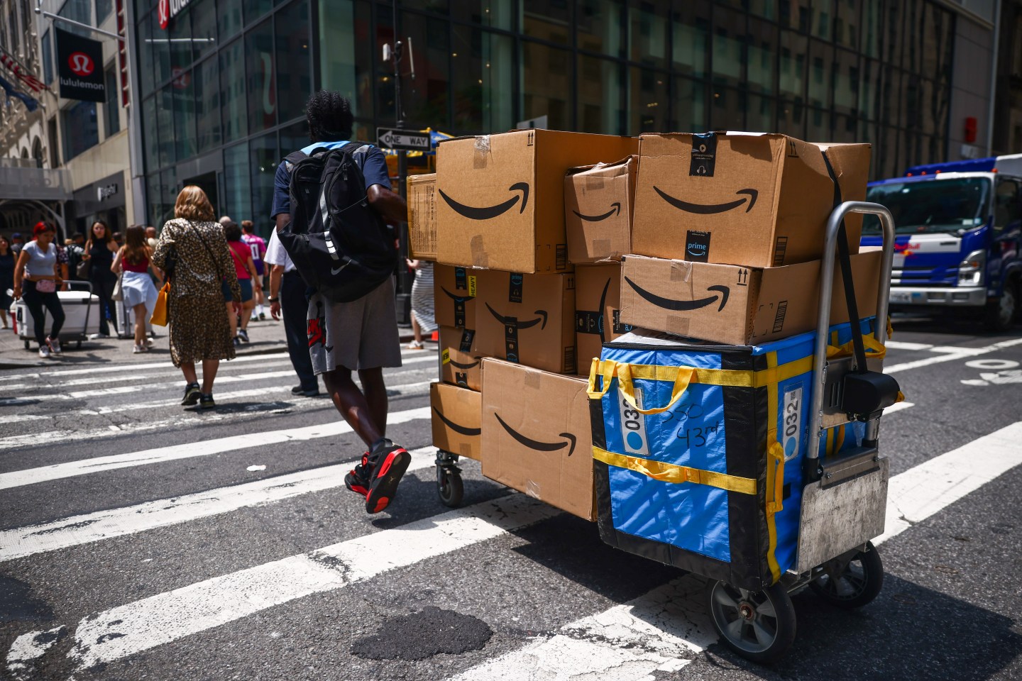 An Amazon delivery person wheels a stack of Amazon boxes across a New York City crosswalk
