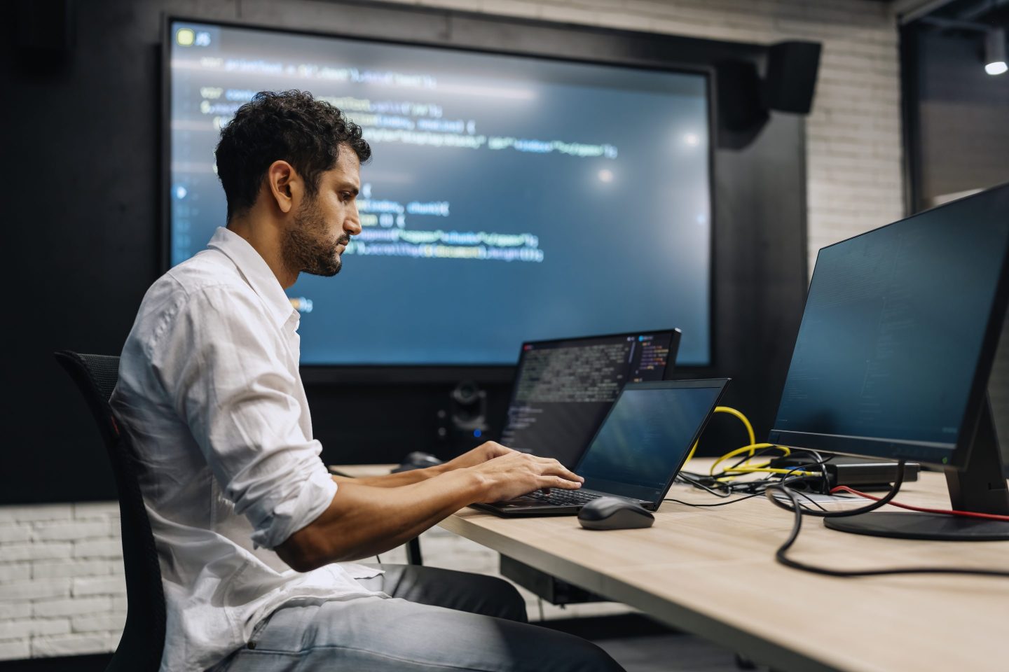 A man works at a table with a laptop on it. A projector in the background displays code.