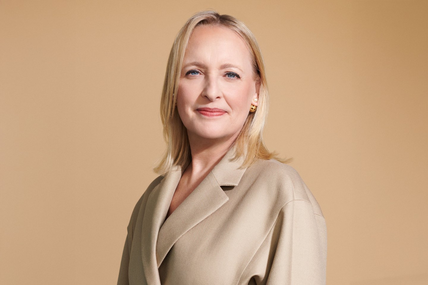 A portrait of Julie Sweet in a beige jacket against a gold backdrop