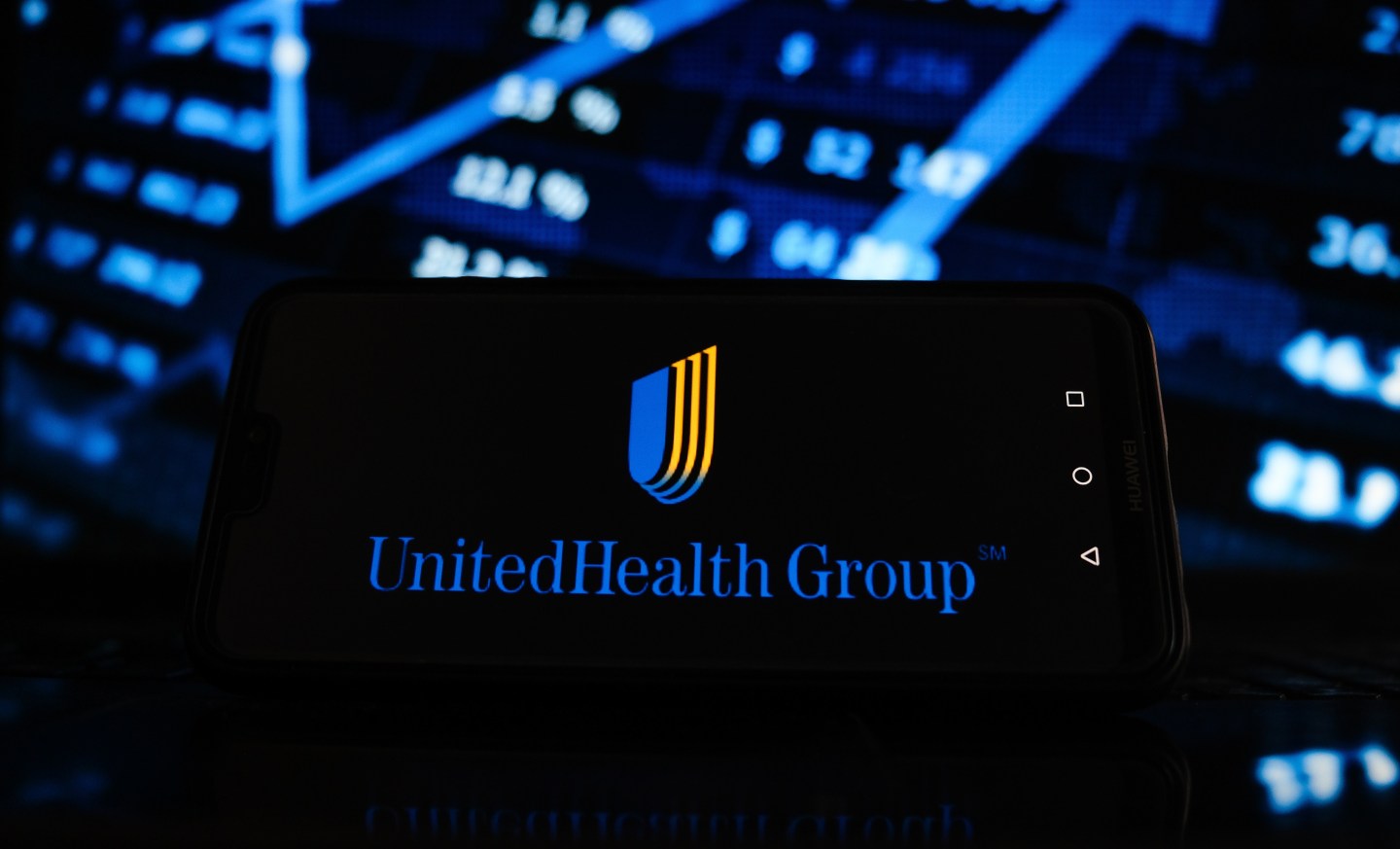 In this photo illustration a United Health Group logo seen displayed on a smartphone screen with stock market in the background.