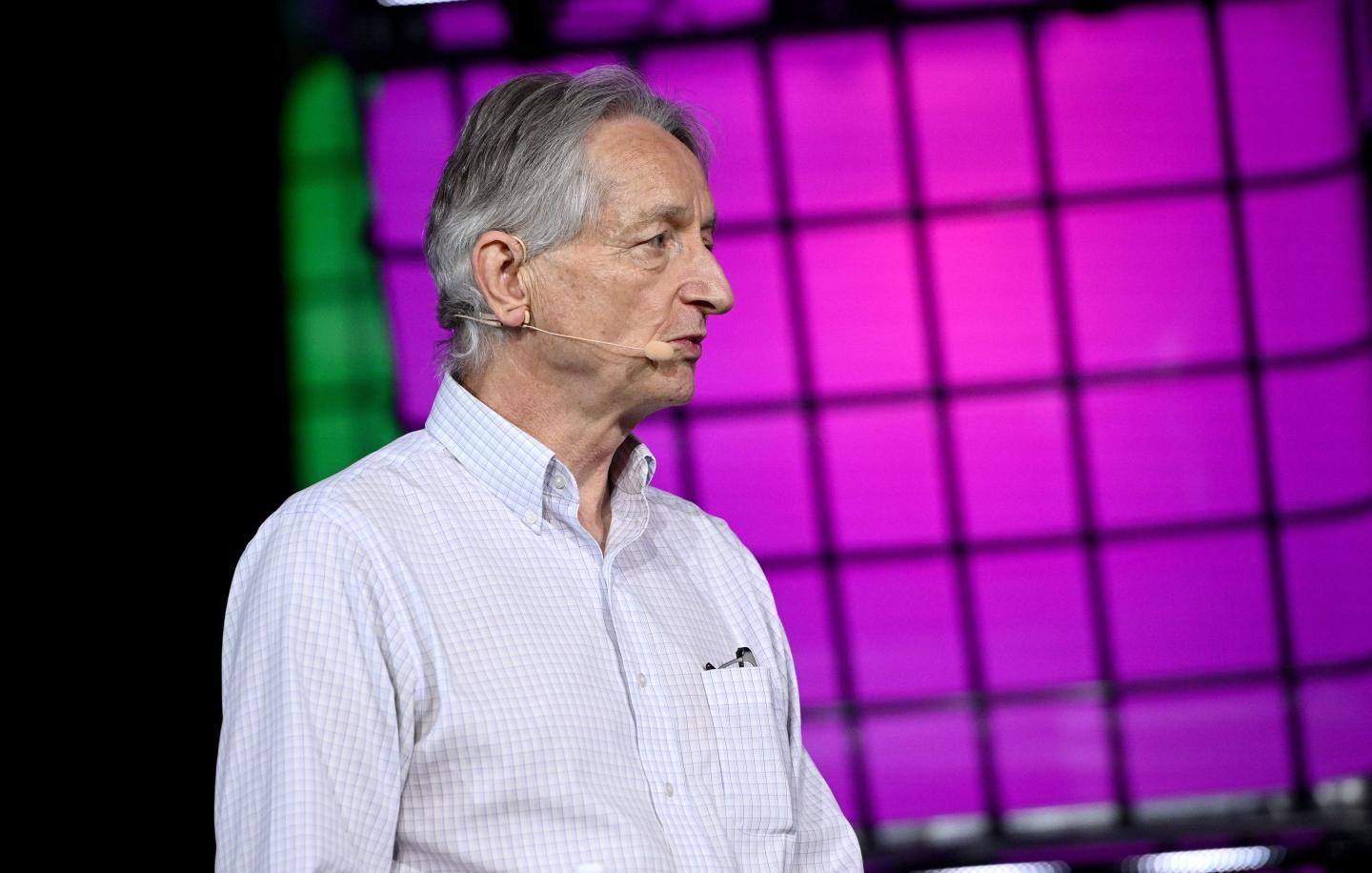 Geoffrey Hinton, stands on stage with a microphone headset. He is in front of a magenta screen.