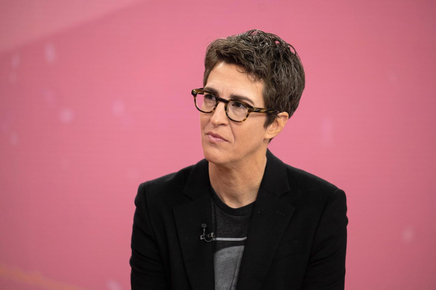 Rachel Maddow looks at an interviewer in front of a pink background.