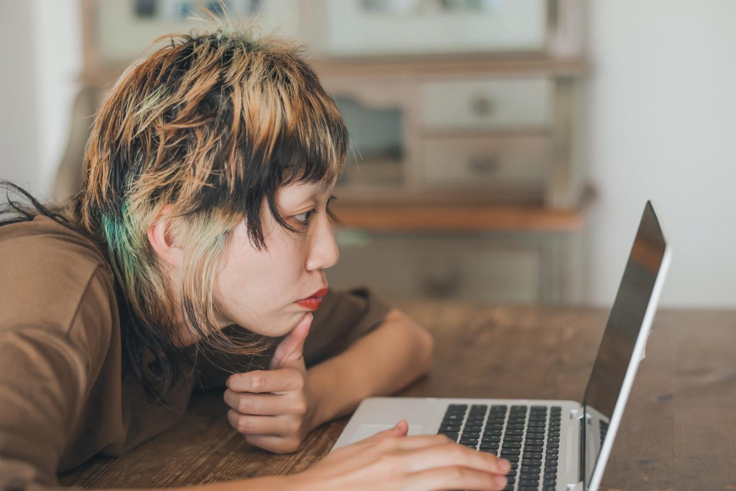 A person with blonde- and blue-streaked hair looks disappointedly at a laptop.