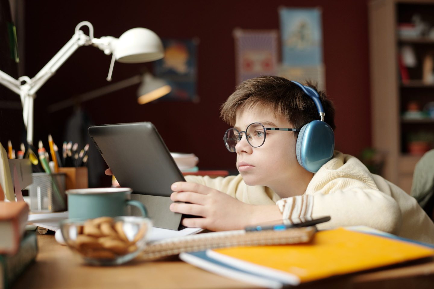 A boy sits at a cluttered desk with headphones on, looking at an iPad.