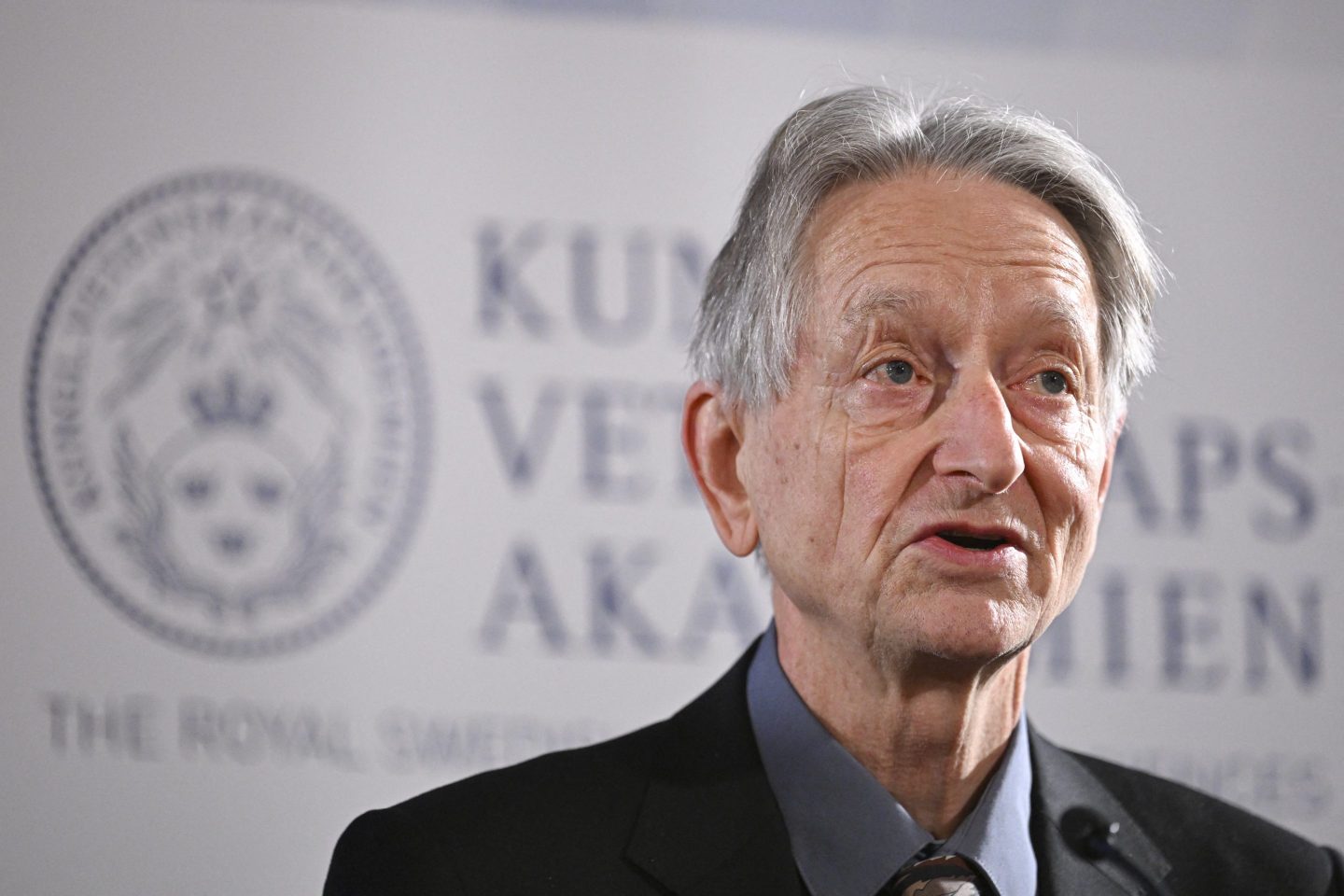 Geoffrey Hinton standing in front of a white and grey background.