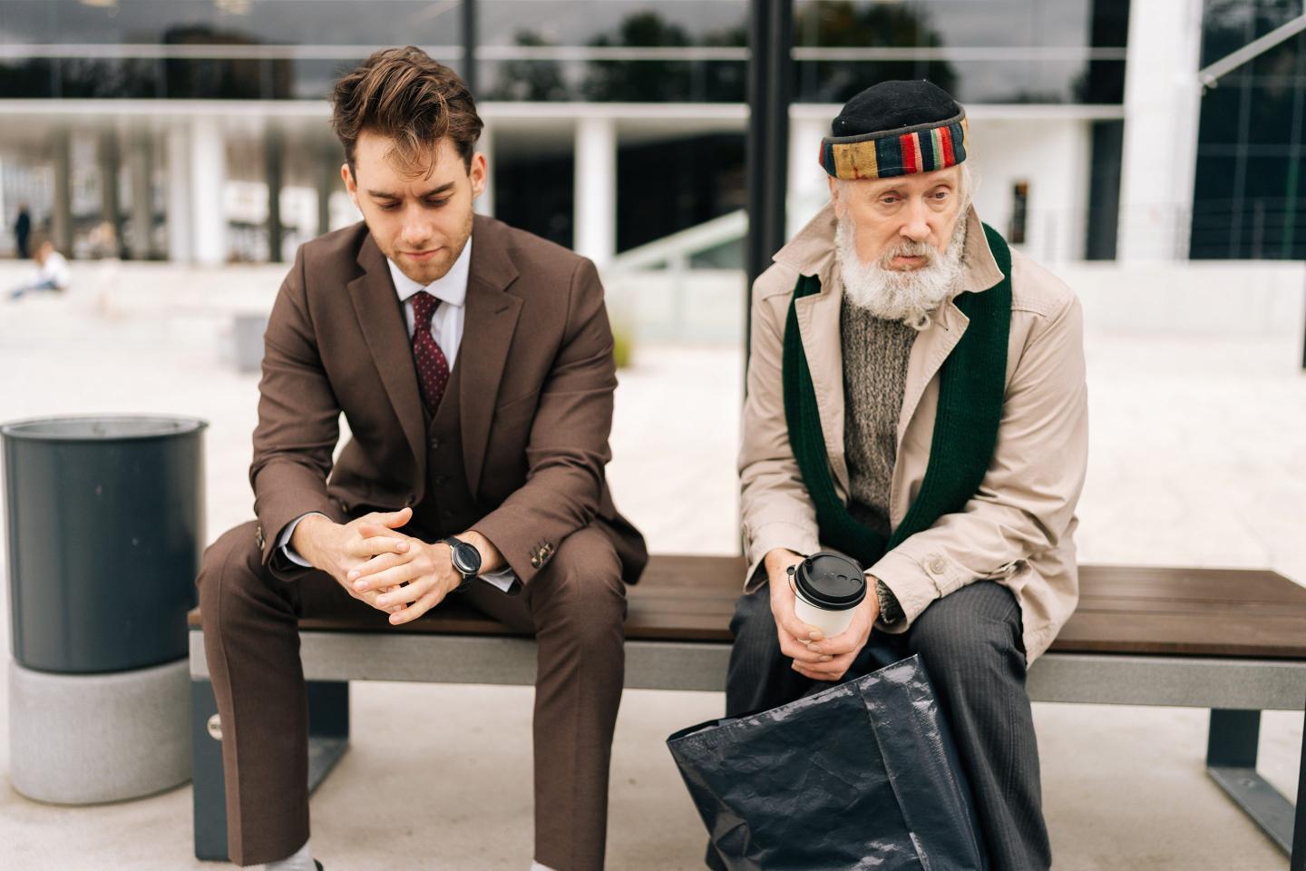 A young man in a suit sits on a bench next to a man who appears less affluent