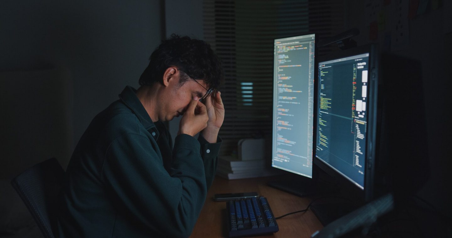 A man sitting in front of a computer screen rubbing his eyes and appearing despondent.