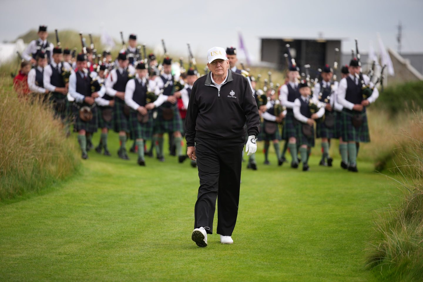 trump walks on a golf course followed by bagpipers
