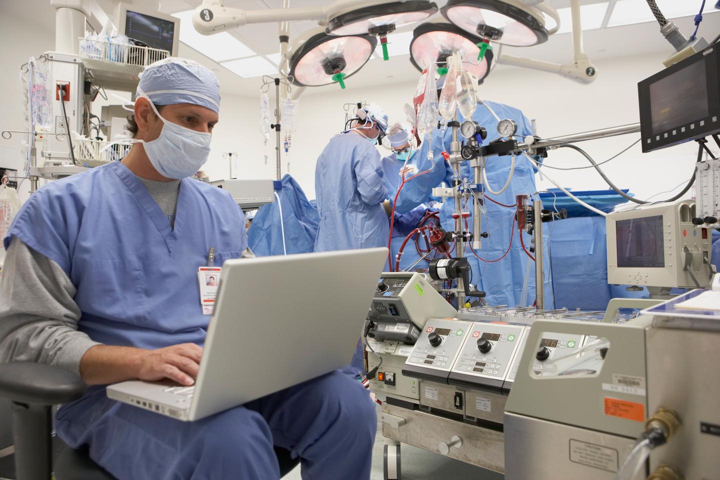 A doctor in an operating room looks at a laptop. Other doctors are gathered in the background.