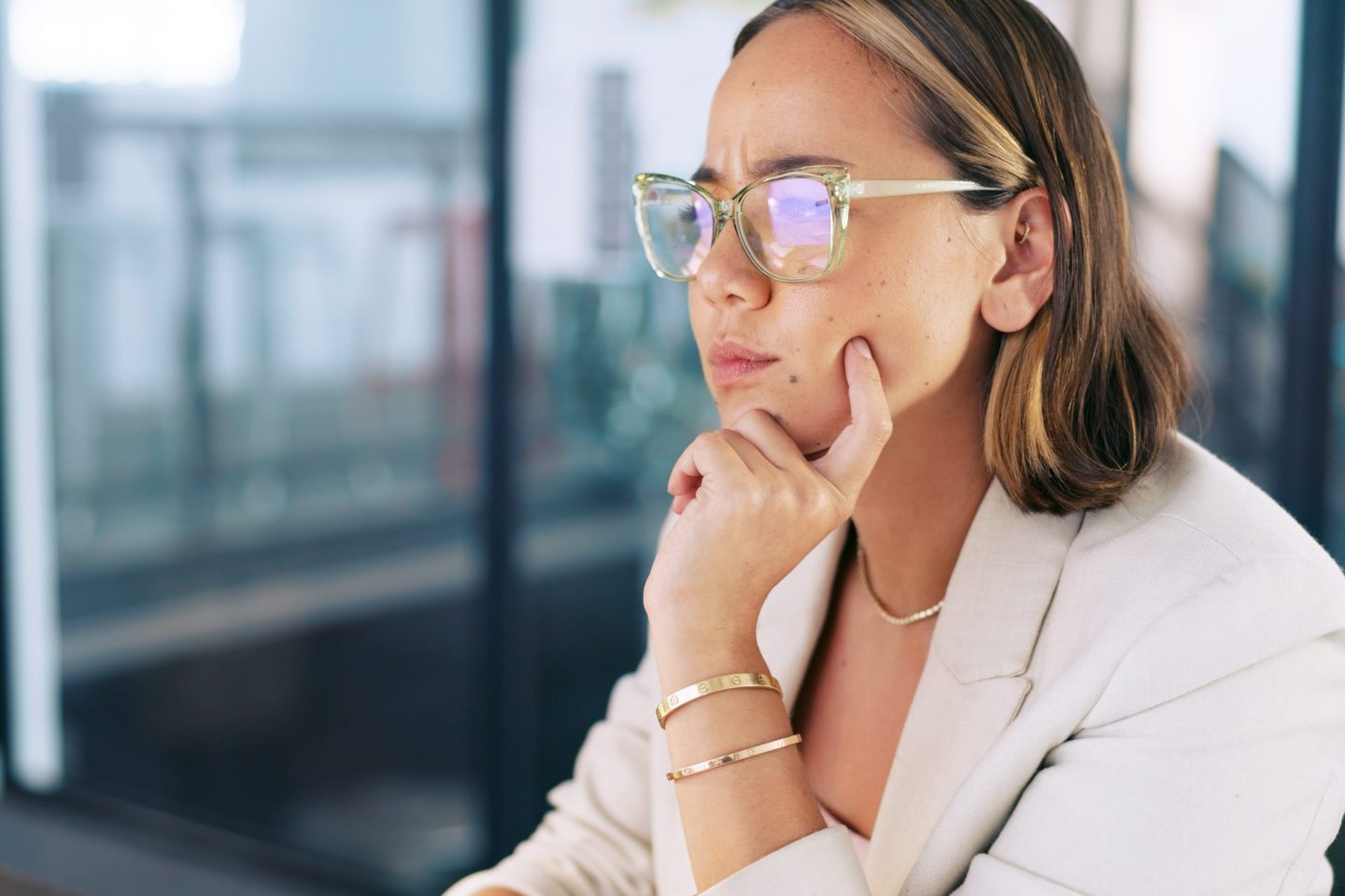A woman wearing glasses and business attire sits at a computer with a confused look on her face