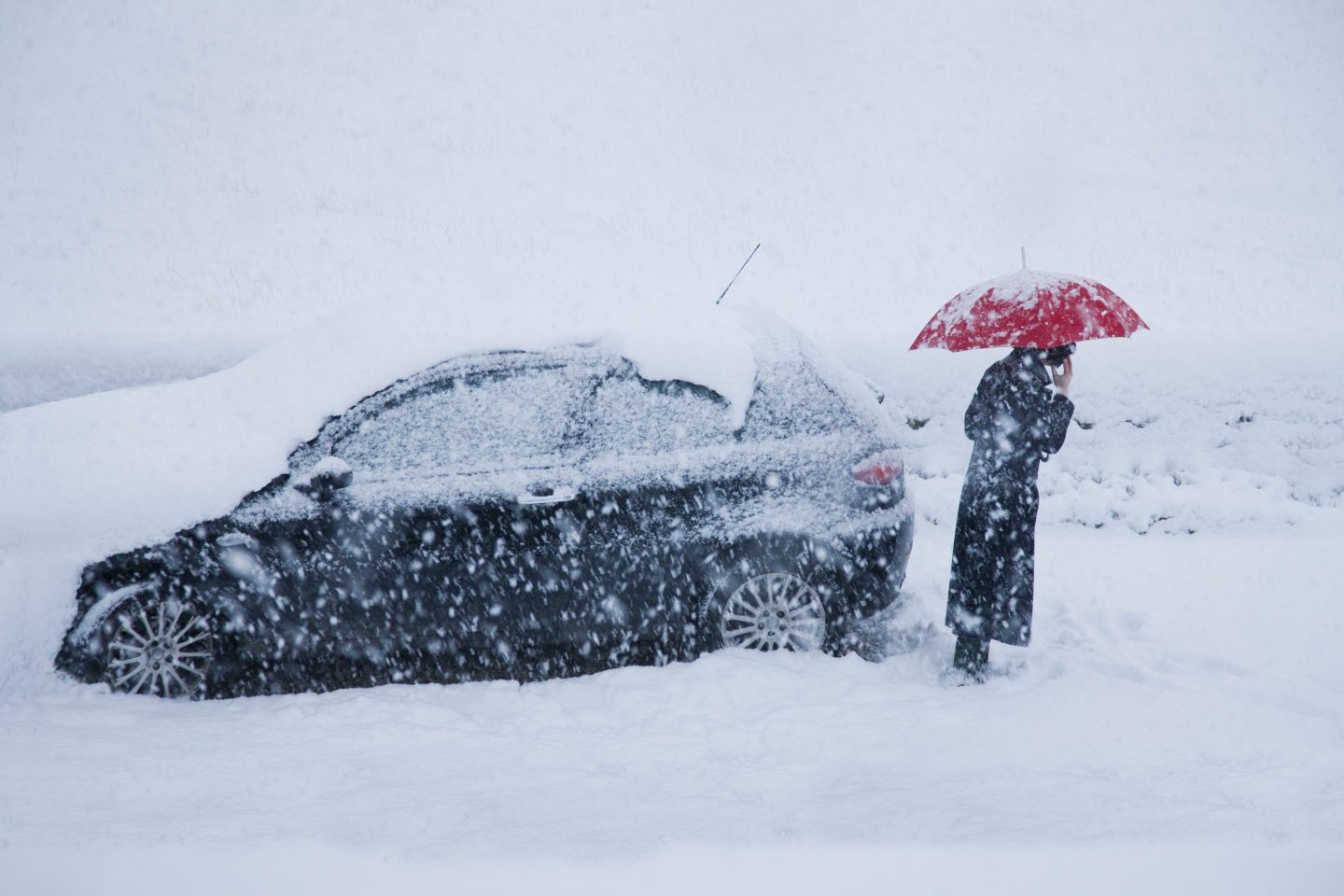 A person with a read umbrella standing by a black car that is stuck in snow in the middle of a blizzard.