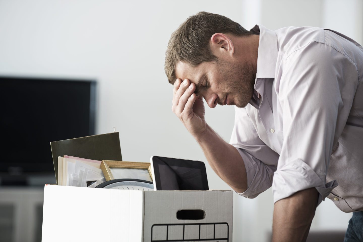 A man with his head in his hand leans over a desk with a box keeping his work posessions.
