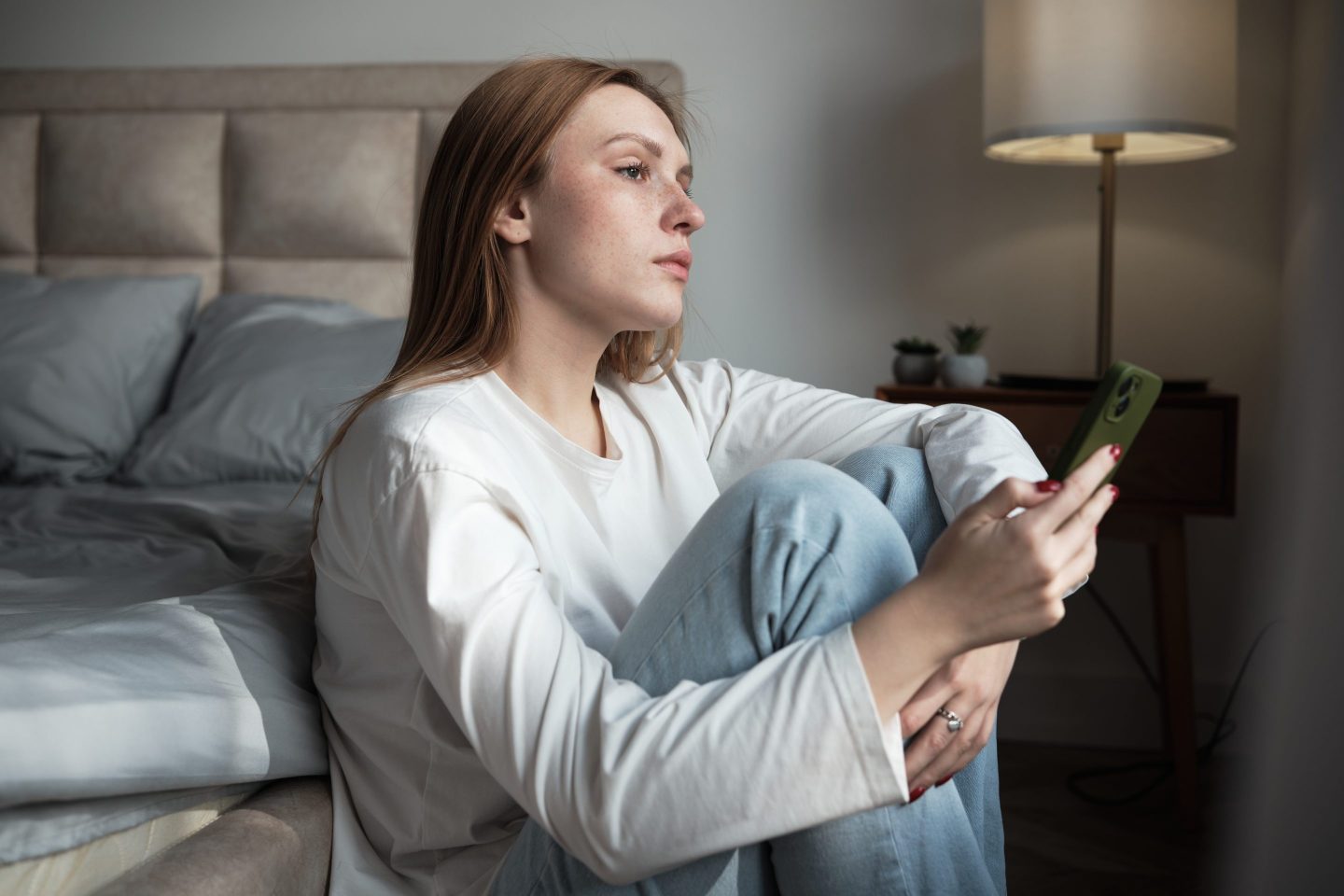 Sad young woman with phone at home, using mobile phone feeling depressed, upset gen z girl holding smartphone looking out window.