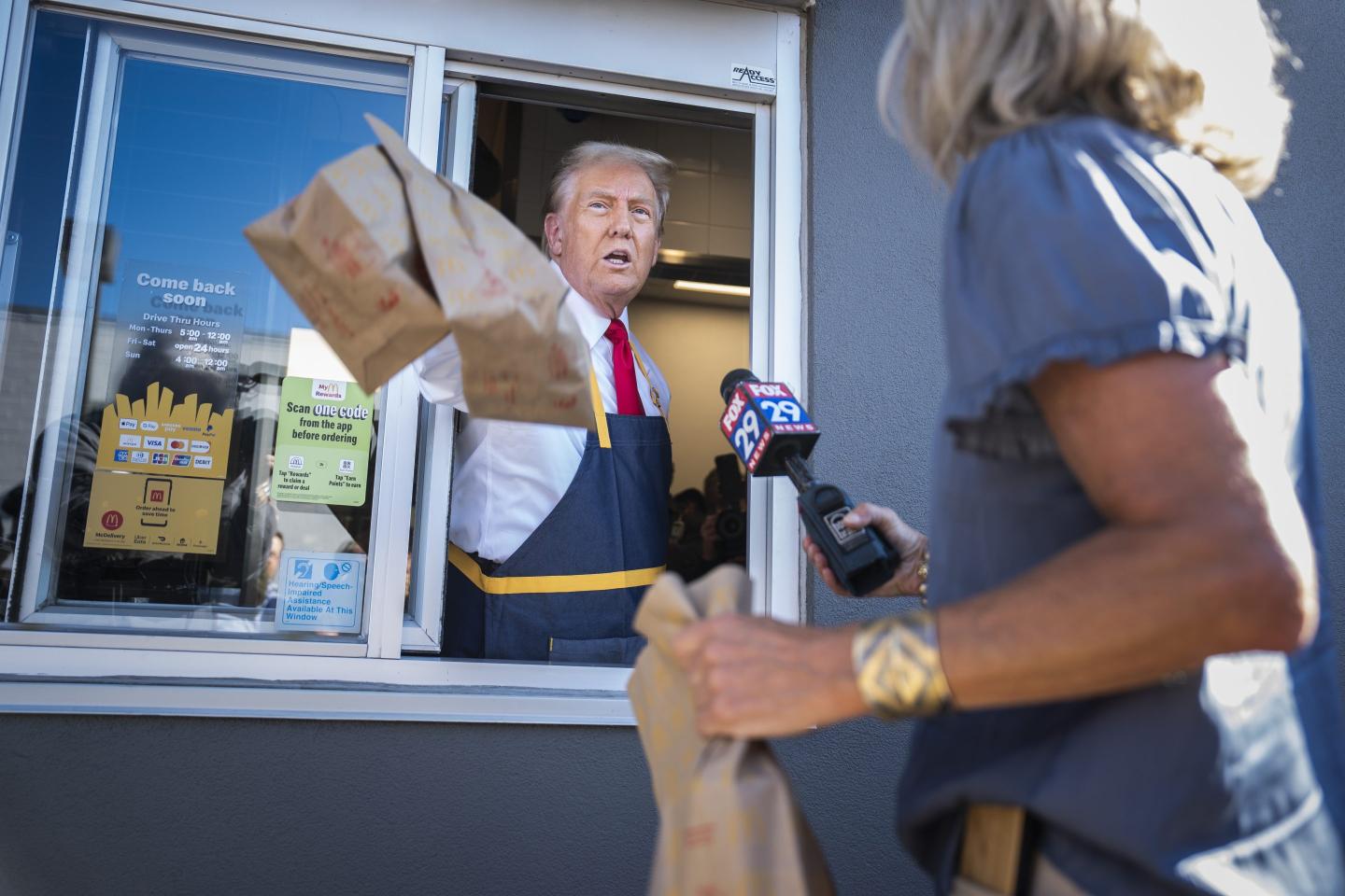 Donald Trump stands in a McDonald's drive-thru window, extending two paper bags in front of him.