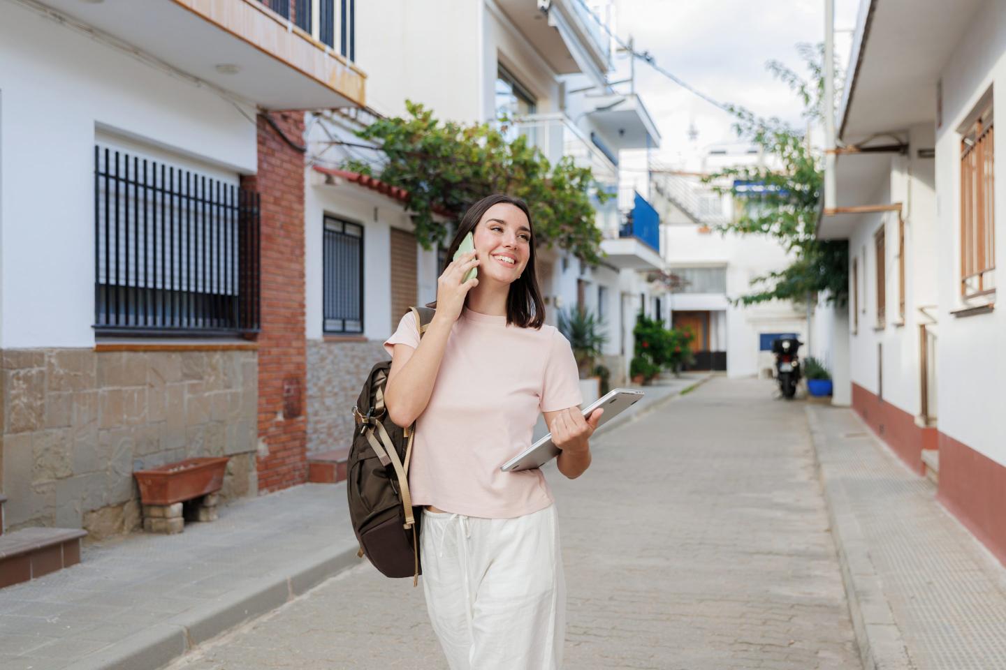 Freelancer woman with backpack and tablet walking down a narrow street in a european town, talking on the phone and smiling